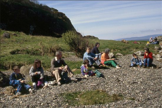 Group of people sitting on a pebble beach eating pack lunches.